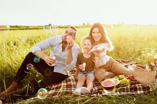 Togetherness Concept. Portrait of cute little daughter blowing soap bubbles with her parents sitting on blanket photo
