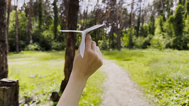 Female hand playin with paper airplane at summer forest. Girl walking with toy plane at woodland on sunny day. Young woman having fun at nature. Concept of leisure outdoor. Close up photo