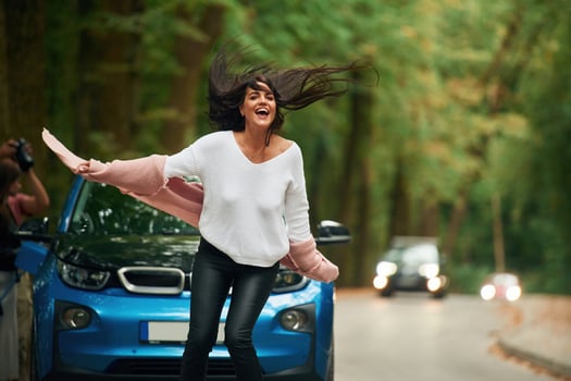 Black automobile behind.. Cheerful beautiful woman is near blue car outdoors photo