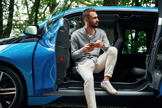 With ,smartphone in hands. Beautiful young man is sitting in the blue car outdoors photo