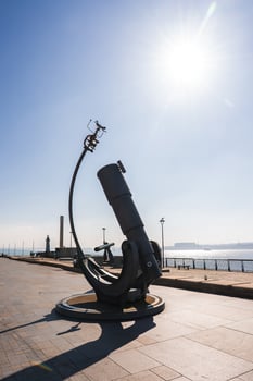 A large telescope like sculpture on a paved promenade in Liverpool, with the River Mersey, industrial structures, and a tall chimney in the background. photo