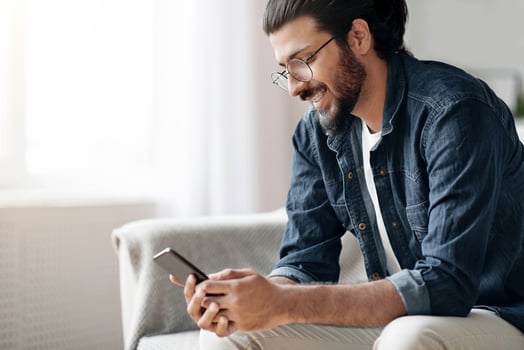New Mobile App. Stylish Western Millennial Guy Using Smatphone While Sitting On Sofa At Home, Browsing Internet, Messaging With Friends, Playing Online Games Or Reading Sms, Wearing Glasses And Braces photo