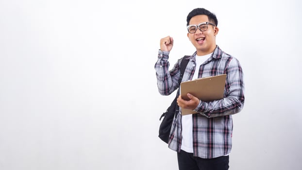 Excited young Asian male college student with a backpack holding wooden clipboard and showing his fist to celebrate victory win success isolated on white background photo