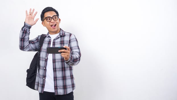 Happy young Asian male college student with a backpack playing smartphone horizontally isolated on white background photo