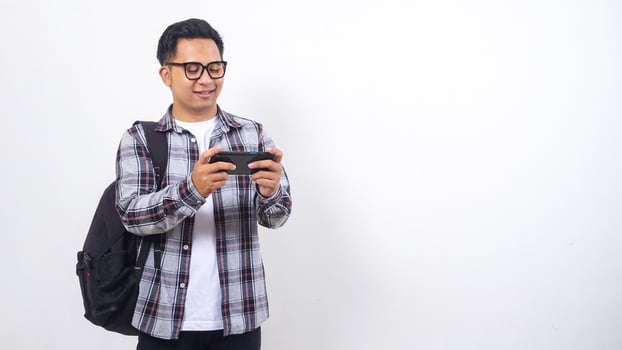 Handsome young Asian male college student with a backpack playing smartphone horizontally isolated on white background photo