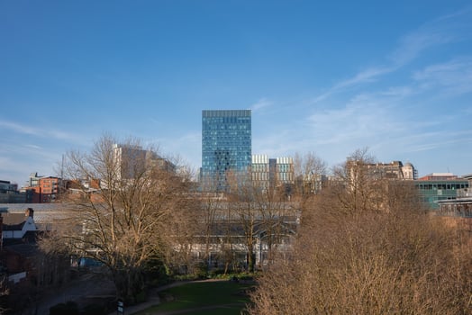 View of Manchester featuring a modern glass skyscraper, historic and contemporary buildings, and a park with bare trees under a clear blue sky. photo