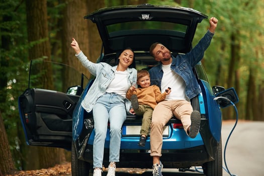 Sitting, on the back of a vehicle. Mother, father and little son are waiting for electric car to charge outdoors photo