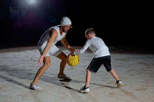 Father and son enjoy a night basketball game at a local park under bright lights focusing on teamwork photo