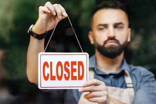 Closure of small businesses, problems in flowers shop during covid-19 quarantine. Sorrowful millennial bearded caucasian man in apron turns nameplate is closed, in store with different plants photo