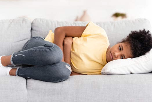 Black Teen Girl Suffering From Stomachache Having Painful Periods Lying Touching Stomach On Couch Indoors. Abdominal Pain Concept photo