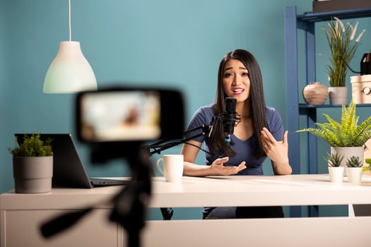 Young female vlogger seated at desk, talking into her podcast mic about marketing strategy for her channel. Asian woman using a camera on tripod, creating lifestyle content for her online audience. photo