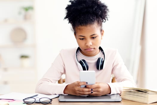 Black Teen Girl Using Mobile Phone Sitting At Laptop Computer Indoors. Home Education And Procrastination photo