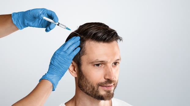 Mesotherapy for hair. Attractive man receiving injections in his head, grey studio background. Man having mesotherapy session at beauty salon, therapist in protective glove with syringe, copy space photo