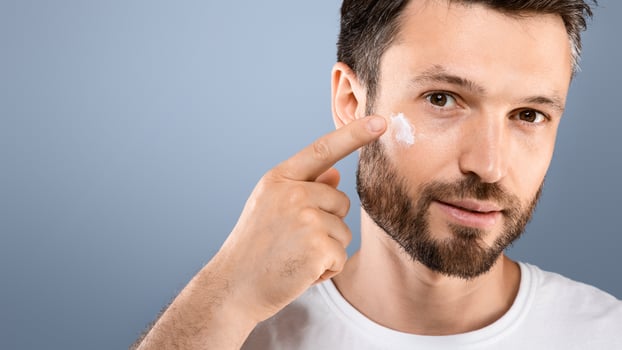 Closeup of bearded handsome man applying face cream on grey studio background, copy space. Good looking middel aged man using beauty products for his face, moisturising or nourishing skin photo