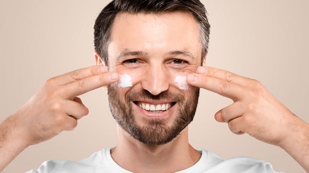 Smiling handsome man applying anti-aging cream on his face, closeup. Middle-aged bearded man with cream on his cheeks from wrinkles and dryness, grey studio background, eye and face care for men photo