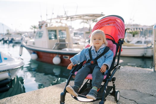 Little smiling girl sits in a stroller on the pier near the moored yachts and points to the sea photo