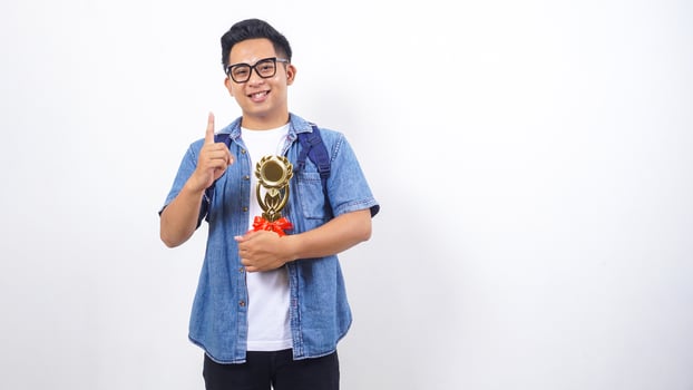 Handsome young Asian male university student in casual outfit with backpack is holding trophy isolated on white background with copy space photo