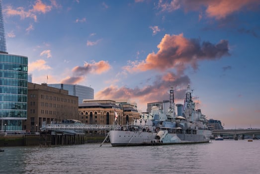 HMS Belfast moored on the River Thames in London, framed by The Shard and city architecture. A vibrant sunset with pink and orange hues lights the scene. photo