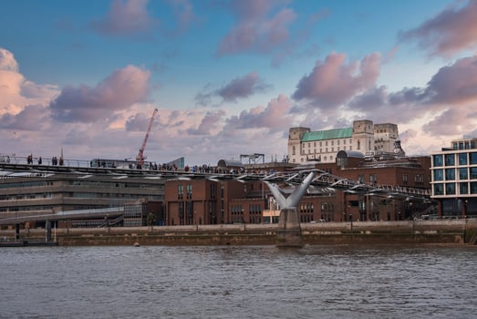 Millennium Bridge in London spans the River Thames, busy with pedestrians. Tate Modern and surrounding architecture are visible under a pink and blue sky. photo