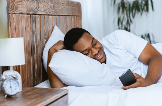 Happy African Man Using Mobile Phone Playing Online Games And Browsing Internet On Smartphone Lying In Bed At Home. Weekend Morning Leisure, Mobile App Concept photo