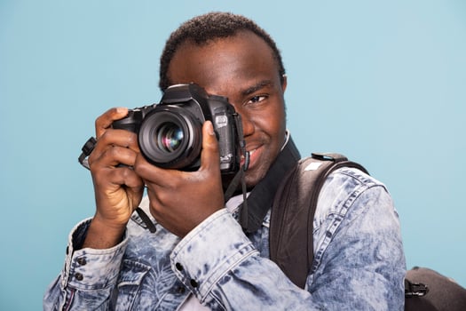 African American male photographer demonstrating the usage of a DSLR. Smiling black man, carrying a backpack, holds a professional camera close to his face as he focuses on his next shot. photo