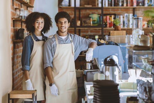 Meeting clients in cafe after covid-19 lockdown. Smiling african american guy and female in aprons and white gloves with espresso machine and takeaway coffee equipment in modern loft cafe interior photo
