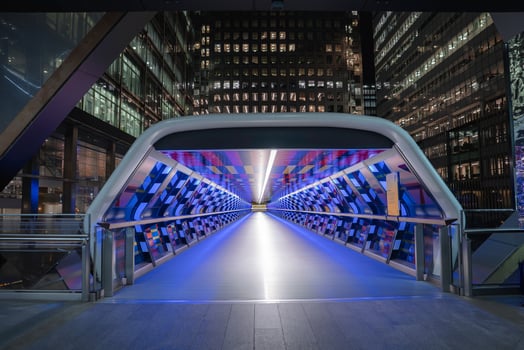 The Crossrail Place Bridge in London glows with blue and purple lights at night, featuring geometric patterns and surrounded by modern office buildings. photo