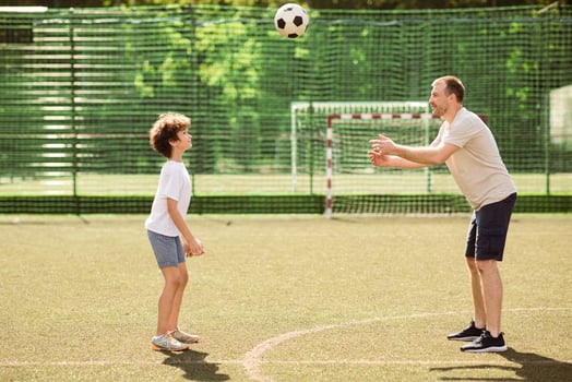 Active Lifestyle Concept. Side view of sportive single adult dad playing football with his son on green grass at soccer pitch, throwing the ball to boy, family spending time together. Recreation photo
