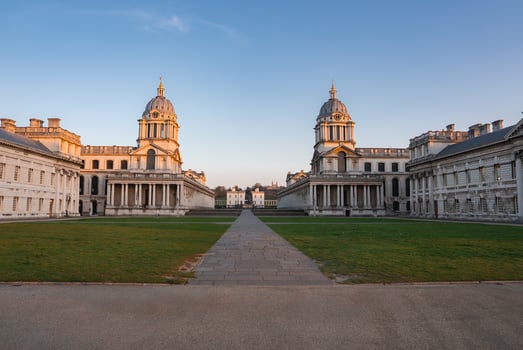 The Old Royal Naval College in Greenwich, London, features two domed buildings flanking a central pathway, with a green lawn and clear blue sky. photo