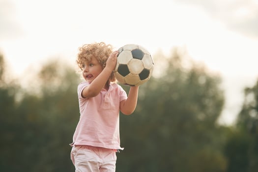 Holding,, soccer ball in hands. Cute little girl with curly hair is playing on the field at daytime photo
