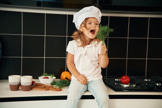 Cheerfu,l little, girl in ,chief hat is sitting on the table of kitchen with vegetables photo