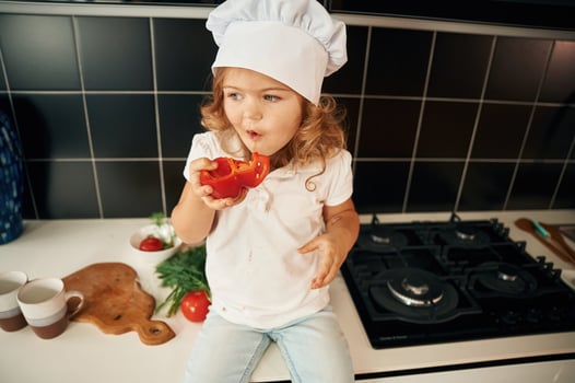 Sitting, and eating pepper. Cheerful little ,girl in chief hat is on the table of kitchen with vegetables photo