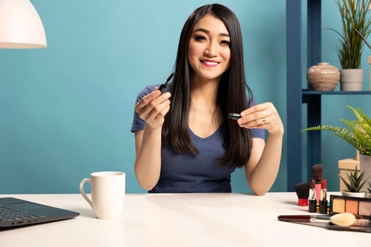 Asian makeup artist with a mascara in her hand, shooting a tutorial for her online audience. Female vlogger sitting at desk and recording a livestream, showing her beauty routine to her fans. photo