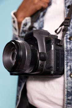 Selective focus on camera worn by black individual in casual outfit, emphasizing design of device. Closeup of DSLR hanging over white shirt and denim jacket, focusing on details of photography gear. photo