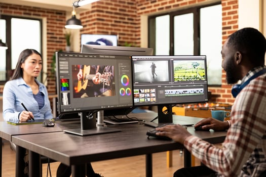 Black media creator operates editing software, merging clips and audio in a diverse production department office. African american man seated at shared creative workstation, working on projects. photo