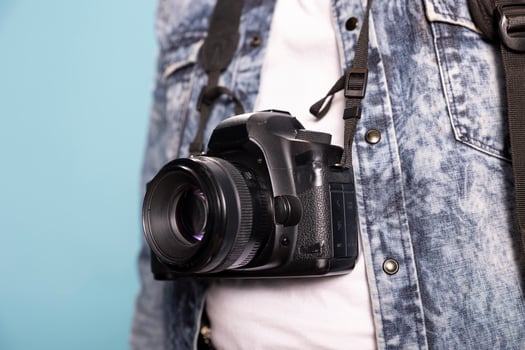 Closeup of a DSLR hanging over an individual wearing a denim jacket and white shirt against an isolated background. Selective focus on a professional camera positioned on the body of a person. photo
