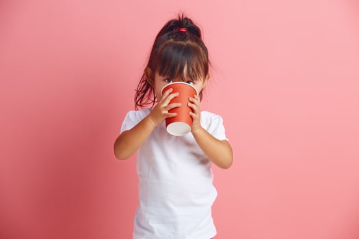 Handsome little asian girl drinking from disposable paper cup standing over pink isolated background. photo