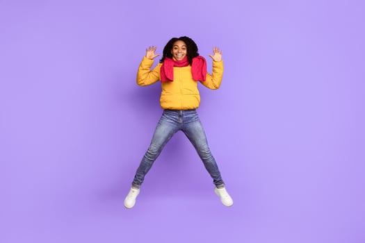 Hello. Joyful African American Woman Jumping Waving Hands In Mid-Air And Smiling To Camera Posing Wearing Warm Jacket Over Blue Studio Background. Winter Advertisement Banner. Full Length Shot photo