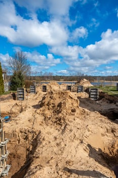 A construction site featuring sand piles, metal framework, and concrete forms. Open land with sparse trees and a distant body of water are visible. photo