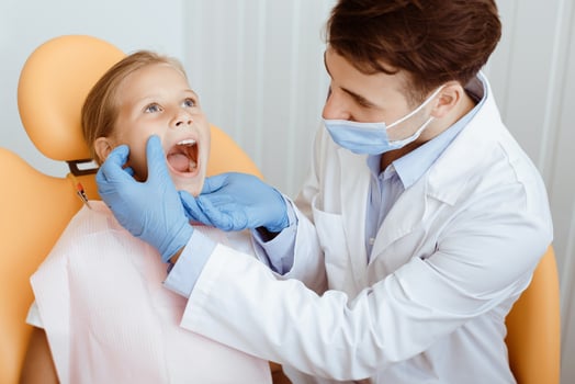 Caries or perfect teeth, tooth care and toothache. Millennial male doctor in white coat, protective mask and gloves examine oral cavity of little girl in chair in modern clinic interior photo
