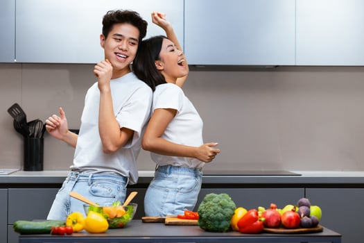 Chinese Spouses Dancing In Kitchen, Having Fun Cooking Together. Couple Enjoying Dinner Preparation At Home. Joyful Family Cooks Meal Indoors. Free Space For Text photo