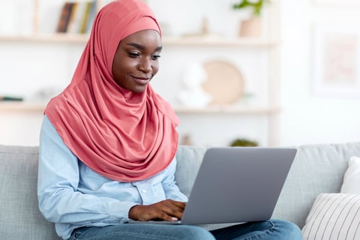 Online Communication. Smiling Black Muslim Woman In Headscarf Using Laptop At Home, African Islamic Lady Messaging With Friends, Typing On Keyboard While Relaxing On Couch In Living Room, Copy Space photo
