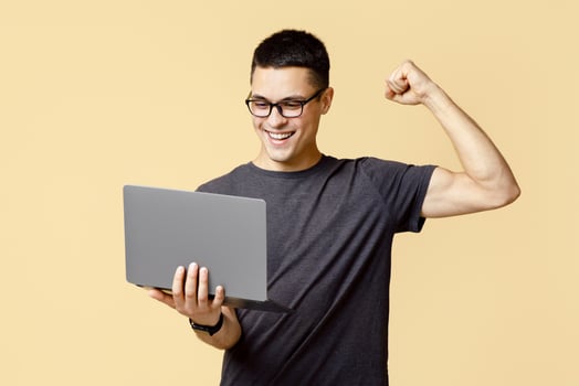 Online win, success at work and great offer. Happy young blogger guy in glasses holds laptop and expresses emotions of victory, isolated on light sand background, empty space, studio shot photo