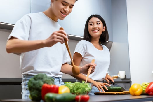 Happy Married Asian Couple Cooking Salad Together Standing Near Table In Modern Kitchen, Husband Helping Wife Cook Meal Preparing Dinner At Home On Weekend. Spouses Enjoying Meal Preparation photo