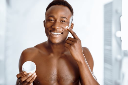 Male Facial Skincare. Cheerful Black Man Applying Cream On Smooth Face Caring For Skin After Shaving Routine, Smiling To Camera Standing Shirtless Near Mirror In Bathroom Indoors. photo