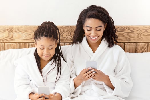 Black little girl and mom in bathrobes sitting with smartphones on bed, paying online games, browsing app, spending time in internet with gadgets, relaxing together at home, shopping online, copy space photo