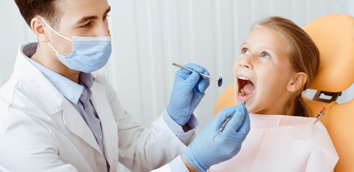 Pediatric dentistry and examination of teeth of small patient. Young male doctor in protective mask in white coat and gloves with medical instruments treats oral cavity of little girl, panorama photo