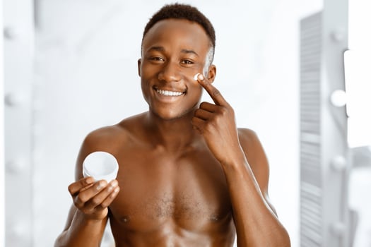 Male Facial Skincare. Cheerful African Guy Applying Cream On Face Holding Moisturizer Jar, Smiling To Camera Standing Shirtless Near Mirror In Bathroom At Home. Male Skin Care And Beauty Concept photo