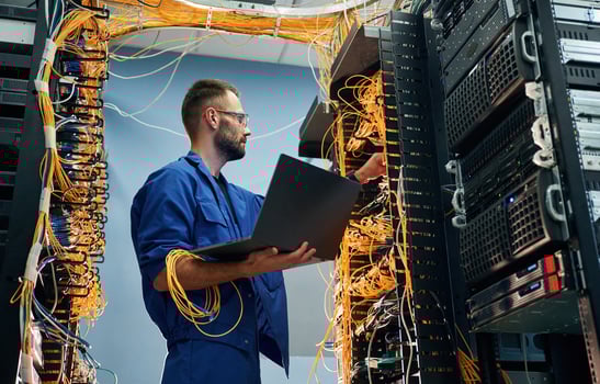 With laptop in hands. Young man, is, working with internet equipment and wires in server room photo