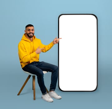 Positive arab guy pointing at big smartphone with white screen while sitting on chair over blue background. Man demonstrating free space for your app or website design, mockup photo